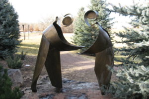 A metal sculpture representing two people, one handing something over to the other, while the other holds their hands out to receive the offering