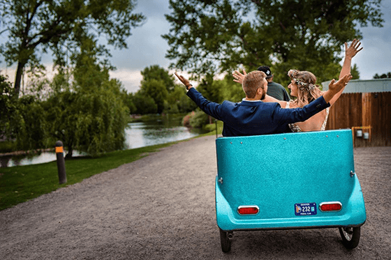A couple in traditional wedding attire, with their arms up in celebration, being driven away from the camera in a turquoise pedicab