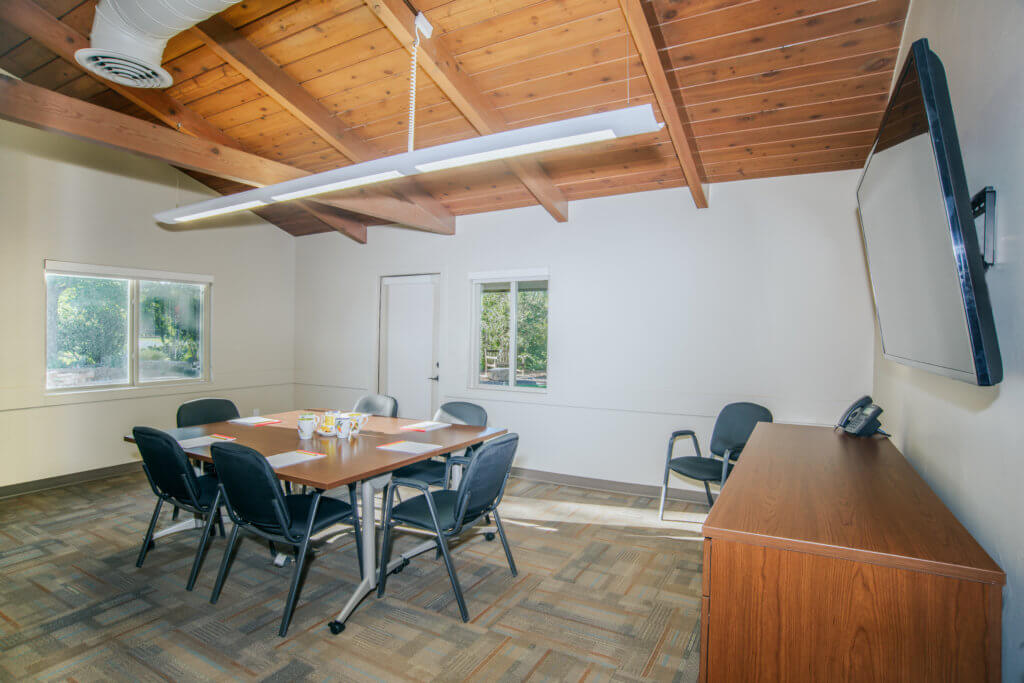 A small room with a square table in the center surtrounded by six office chairs. Another chair sits in the corner and on the right-hand wall is a TV, a credenza and on top on the credenza is a phone