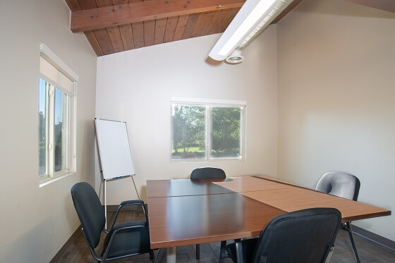 A small room with a square tyble surrounded by 4 office chairs. In the corner is an easel holding an oversized pad of paper