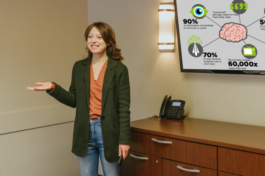 A woman in a green blazer and jeans standing in front of a TV at the Hudson Gardens Residence, giving a presentation