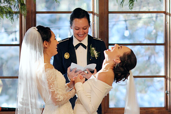 Two women in wedding dresses at their ceremony at the Inn. The women and the person officiating the wedding are all laughing