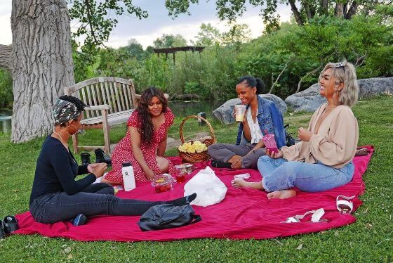 4 women, all sitting on a large red blanket, enjoying a picnic