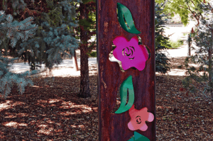 A steel metyal sculpture, that has weathered to be brown, containing brightly painted pink steel flowers and green leaves