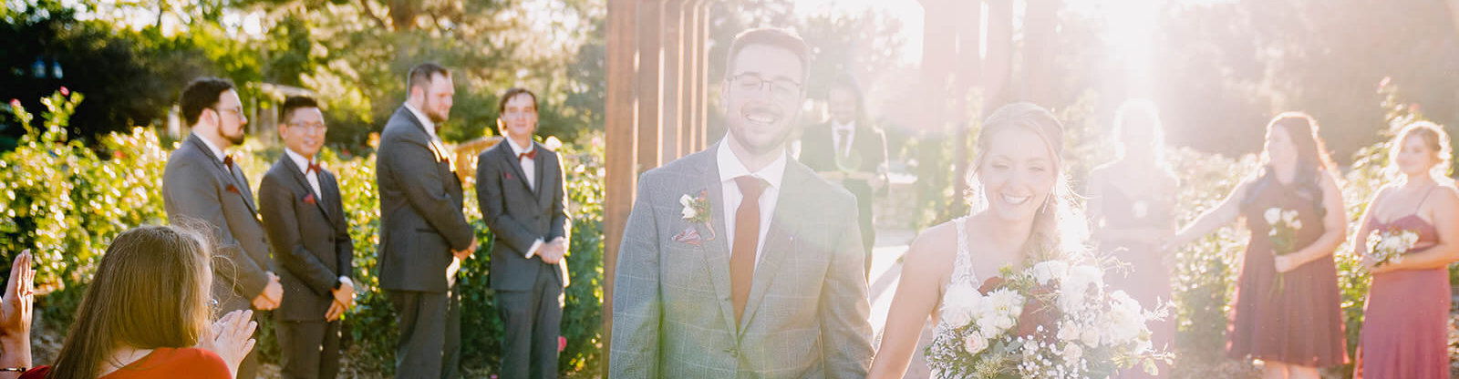 A man and a woman in wedding attire walking away from the rose garden while their wedding party looks on